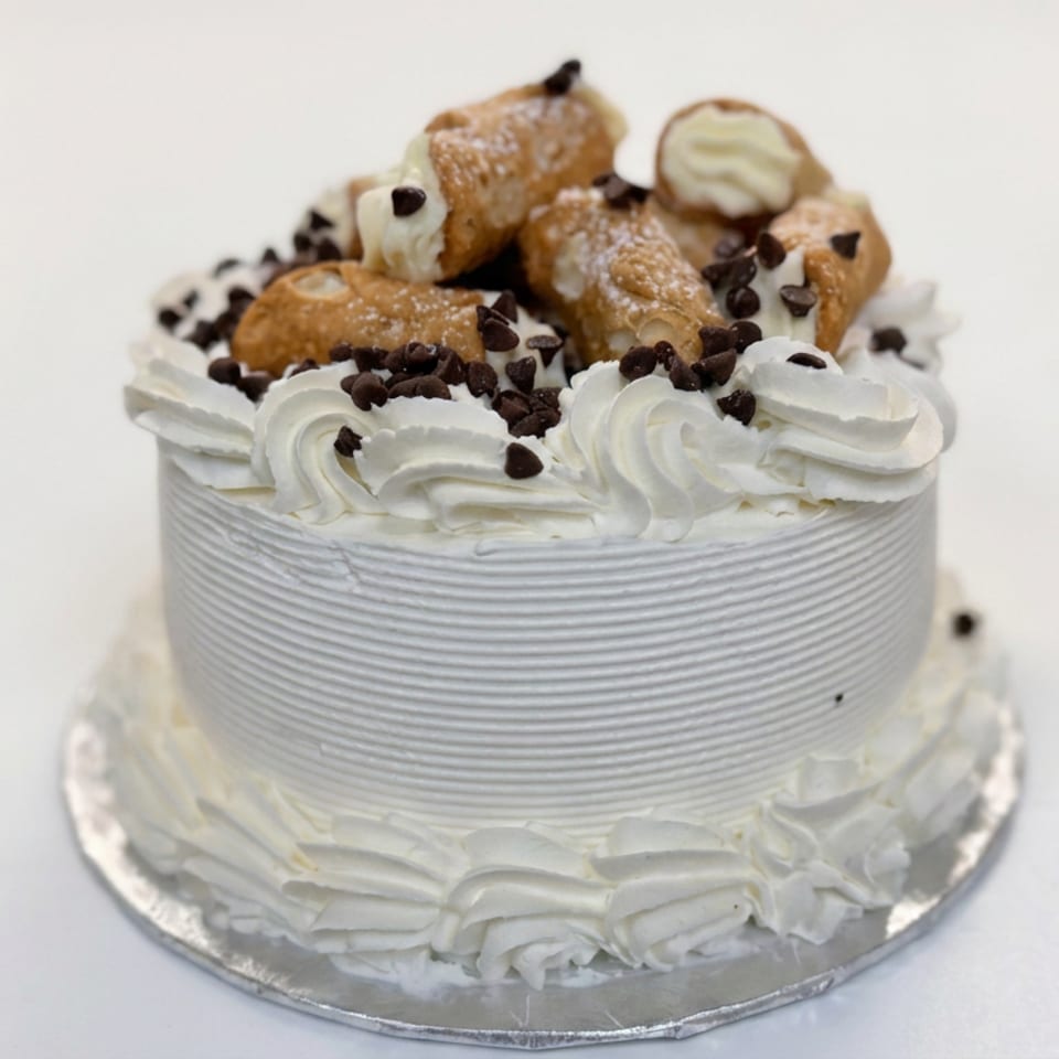 Round white-frosted cake topped with whipped cream, chocolate chips, and cannoli pieces on a silver cake board.