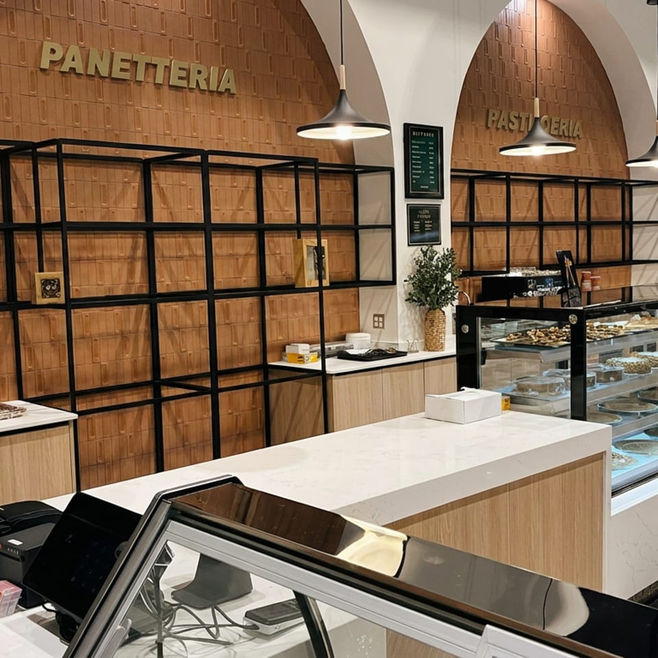 Warmly lit Italian panetteria and pasticceria interior with pastry display cases, shelving, pendant lights, and marble counters.
