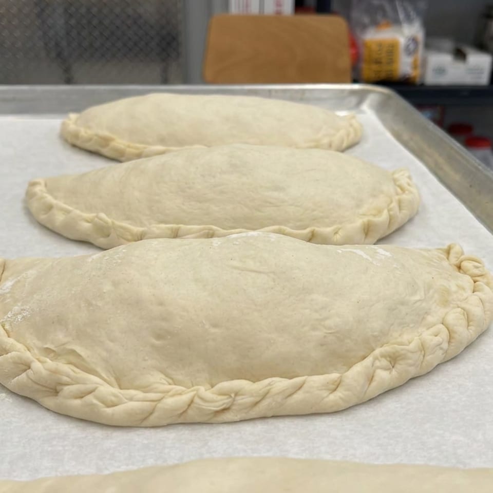 Four uncooked, hand-crimped stuffed dough pastries arranged on a parchment-lined baking tray in a commercial kitchen.